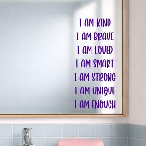 May include: A bathroom mirror with the words "I AM KIND, I AM BRAVE, I AM LOVED, I AM SMART, I AM STRONG, I AM UNIQUE, I AM ENOUGH" in purple lettering. A pink cosmetic bag and a folded purple towel sit on the counter.