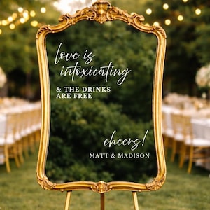 May include: A gold-framed mirror on a gold easel, with the text "love is intoxicating & the drinks are free cheers! MATT & MADISON". The mirror is surrounded by white flowers and greenery, with tables and chairs in the background.