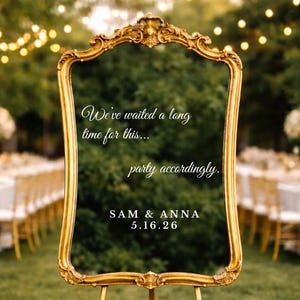May include: A gold-framed mirror on a gold easel, with the text "We've waited a long time for this... party accordingly. SAM & ANNA 5.16.26." The mirror is surrounded by white flowers and set on a grassy lawn.
