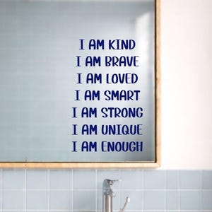 May include: A mirror with the words "I AM KIND, I AM BRAVE, I AM LOVED, I AM SMART, I AM STRONG, I AM UNIQUE, I AM ENOUGH" in blue lettering. The mirror is above a white countertop with two sinks and a chrome faucet.