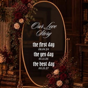 May include: An oval floor mirror with a gold frame featuring the white text Our Love Story, the first day 08.04.24, the yes day 01.11.26, and the best day 03.15.27. The mirror is decorated with dark red and cream roses and green foliage.