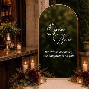 May include: An arched dark green sign with a gold frame featuring the white text Open Bar and the drinks are on us, the hangover is on you. The sign sits on a round table covered with a beige cloth, decorated with dark red flowers and candles.