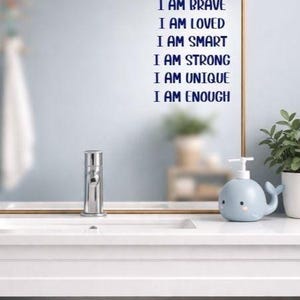Puede incluir: Interior de ba&ntilde;o con un espejo con marco dorado que muestra el texto "I AM KIND, I AM BRAVE, I AM LOVED, I AM SMART, I AM STRONG, I AM UNIQUE, I AM ENOUGH" en azul. Un dispensador de jab&oacute;n azul con forma de ballena est&aacute; sobre la encimera blanca.