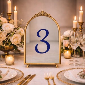May include: An arched table number sign with a gold metal frame and a decorative filigree top. The center of the sign features the number 3 printed in a bold, dark blue serif font. The sign stands upright on a flat surface with small metal feet.