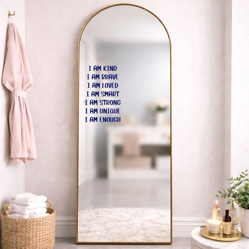 Puede incluir: Un espejo arqueado de cuerpo entero con marco dorado. El espejo tiene texto azul que dice: "I AM KIND, I AM BRAVE, I AM LOVED, I AM SMART, I AM STRONG, I AM UNIQUE, I AM ENOUGH." Una bata rosa cuelga a la izquierda.