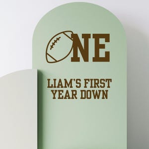 May include: A light green sign with the text "ONE" and a football graphic. Below, the sign reads "LIAM'S FIRST YEAR DOWN" in brown lettering. The sign has a rounded top and a white surface on the left.