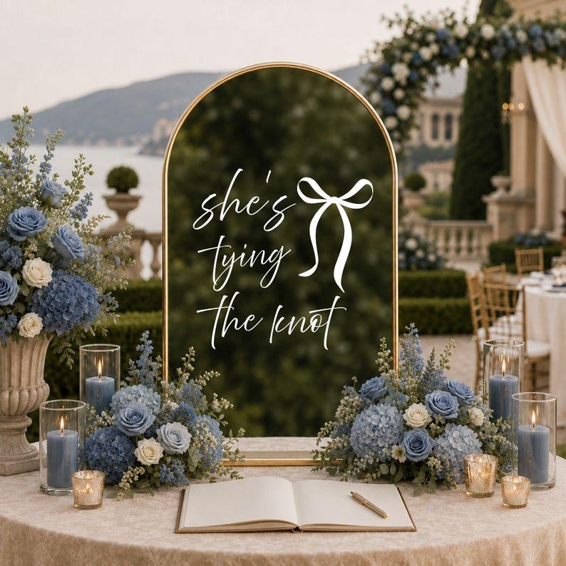 May include: An arched gold-framed mirror sign with the white text "she's tying the knot" and a bow graphic. The sign is displayed on a table with two blue floral arrangements, two glass candle holders with blue candles, and an open guest book.