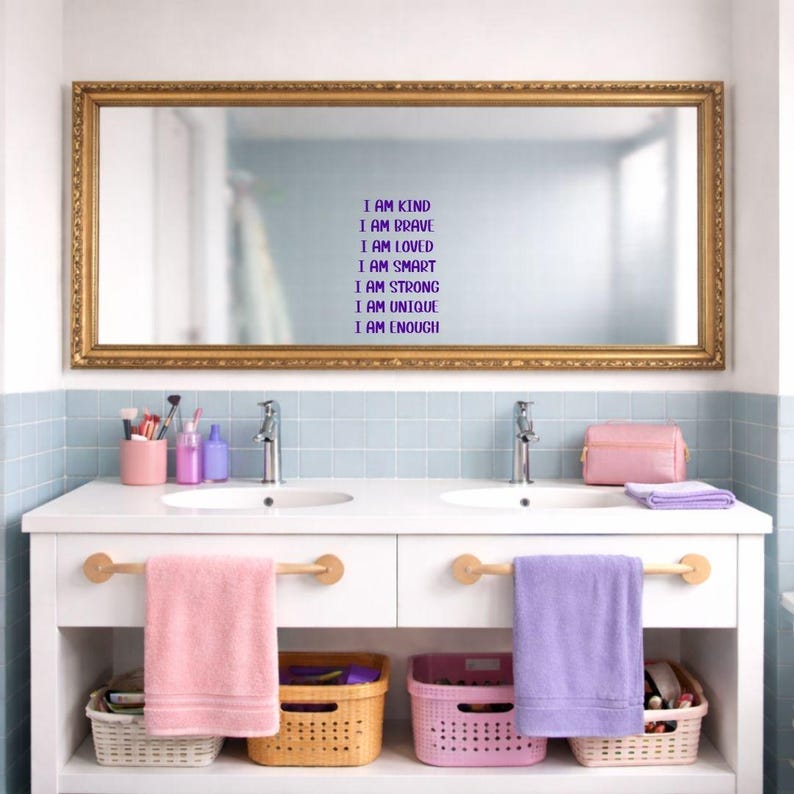Puede incluir: Un mueble de ba&ntilde;o con un gran espejo con marco dorado. El espejo tiene el texto "I AM KIND, I AM BRAVE, I AM LOVED, I AM SMART, I AM STRONG, I AM UNIQUE, I AM ENOUGH" en morado. Debajo hay dos lavabos, toallas rosas y moradas y cestas.