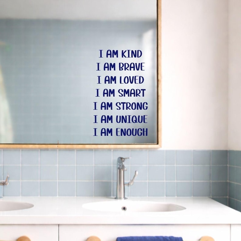 Puede incluir: Un espejo con las palabras "I AM KIND, I AM BRAVE, I AM LOVED, I AM SMART, I AM STRONG, I AM UNIQUE, I AM ENOUGH" en letras azules. El espejo est&aacute; sobre una encimera blanca con dos lavabos y un grifo cromado.