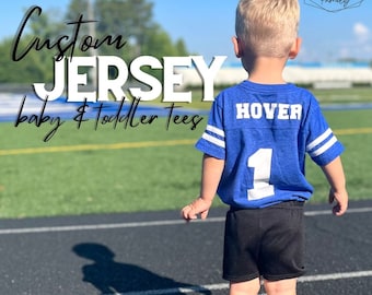 May include: A blue short-sleeved jersey with white stripes on the sleeves, the name HOVER, and the number 1 printed in white on the back. The jersey is paired with black athletic shorts. Text on the image reads Custom JERSEY baby & Toddler Tees.