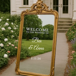 Pu&ograve; includere: Uno specchio da terra alto e rettangolare con una cornice dorata ornata da volute decorative sulla parte superiore. La superficie in vetro mostra un testo bianco che recita: WELCOME TO THE WEDDING OF Ben & Anna 2.15.26, riflettendo l'ambiente esterno.