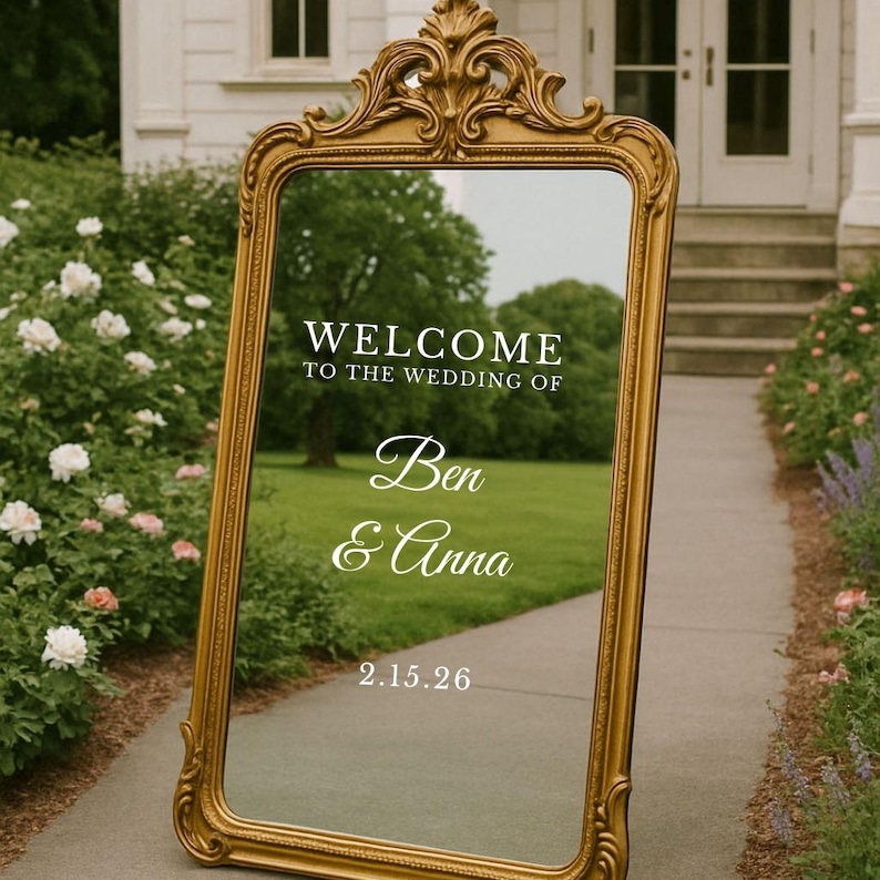 Pu&ograve; includere: Uno specchio da terra alto e rettangolare con una cornice dorata ornata da volute decorative sulla parte superiore. La superficie in vetro mostra un testo bianco che recita: WELCOME TO THE WEDDING OF Ben & Anna 2.15.26, riflettendo l'ambiente esterno.