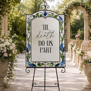 May include: A rectangular sign with a stained glass border featuring green leaves and blue flower accents. The center is a white panel with the black text 'TIL death DO US PART' in a mix of script and serif fonts, supported by a black metal stand.