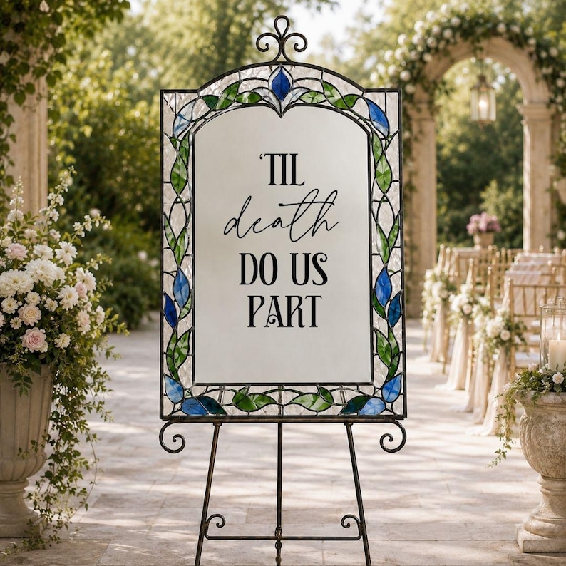 May include: A rectangular sign with a stained glass border featuring green leaves and blue flower accents. The center is a white panel with the black text 'TIL death DO US PART' in a mix of script and serif fonts, supported by a black metal stand.