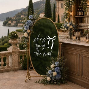 May include: An oval floor mirror with a gold metal frame, featuring white text that reads "she's tying the knot" next to a white bow graphic. The mirror is decorated with arrangements of blue and white flowers and green foliage on the top left and bottom right corners.