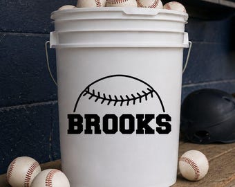 May include: A white plastic bucket with a metal handle, filled with baseballs. The front of the bucket features a black graphic of the top half of a baseball with the name BROOKS printed in bold, capital, black block letters underneath it.