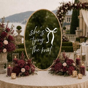 May include: An oval mirror with a gold frame featuring the white text "she's tying the knot" and a bow graphic. The mirror is displayed on a table with burgundy floral arrangements and cylindrical glass candle holders containing dark red candles.
