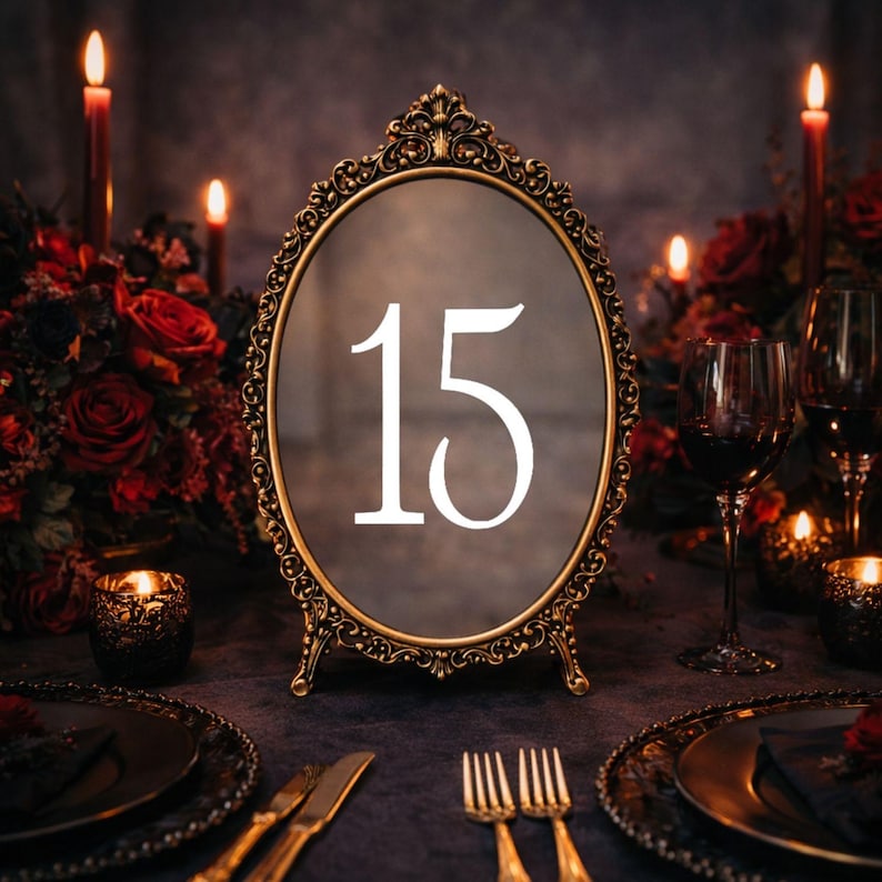 May include: An oval table number sign featuring the number 15 in white serif font centered on a dark reflective surface, encased in an ornate, antique gold-colored metal frame with intricate scrollwork details and a small stand base.
