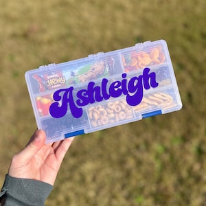 May include: A clear plastic storage container with multiple compartments filled with various snacks. The container has a blue latch and a purple vinyl sticker with the name "Ashleigh" in a retro font.