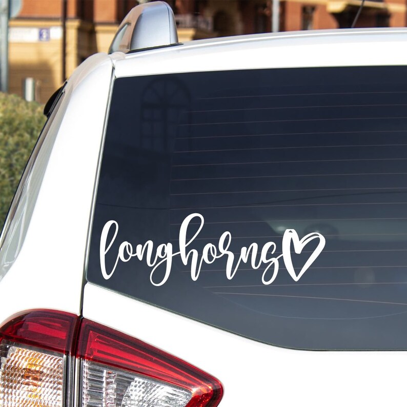 May include: White vinyl decal with the word "longhorns" in cursive lettering and a heart symbol. The decal is on the back window of a white car.