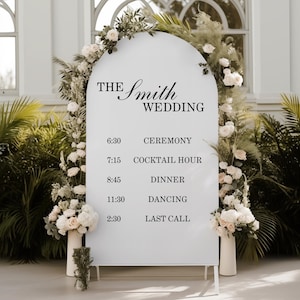 May include: A white wedding sign with an arched top, displaying the text "THE Smith WEDDING" and a schedule. The sign is decorated with floral arrangements and greenery. The schedule includes times for the ceremony, cocktail hour, dinner, dancing, and last call.