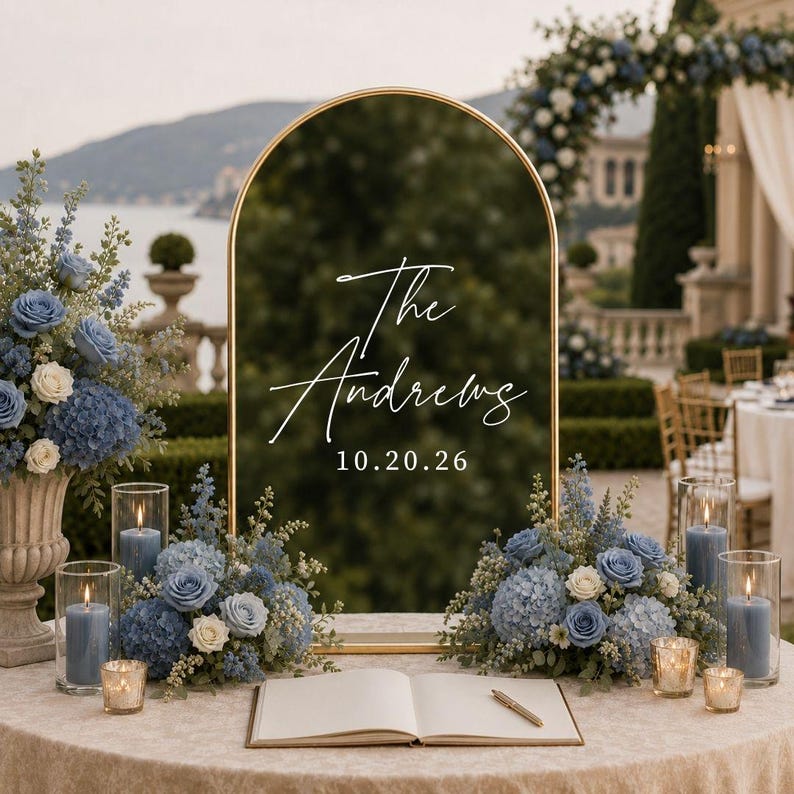 May include: An arched gold-framed mirror sign featuring the white text The Andrews and 10.20.26. The sign is displayed on a table with an open guestbook, blue floral arrangements, and four glass pillar candles with blue wax on a beige tablecloth.