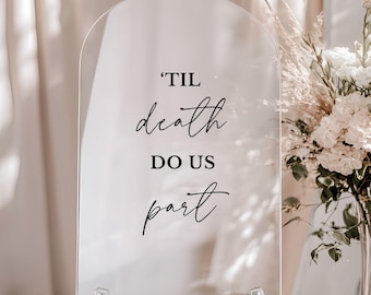 May include: A clear, arched acrylic sign featuring black text that reads 'TIL death DO US part. The sign is supported by a silver metal stand at the base, with the text displayed in a combination of bold block lettering and elegant script.