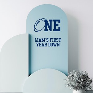 May include: A light blue decorative sign with the word "ONE" and a football graphic. Below, the text reads "LIAM'S FIRST YEAR DOWN" in navy blue. The sign is set against a white background with decorative arches.