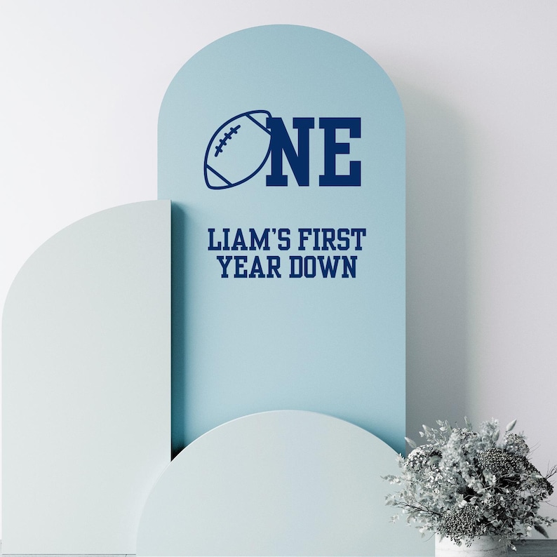 May include: A light blue decorative sign with the word "ONE" and a football graphic. Below, the text reads "LIAM'S FIRST YEAR DOWN" in navy blue. The sign is set against a white background with decorative arches.