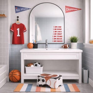 Puede incluir: Un ba&ntilde;o con un tocador blanco, espejo y decoraci&oacute;n deportiva. Una camiseta roja con el n&uacute;mero 1 cuelga de la pared. El espejo tiene las palabras "I AM KIND, BRAVE, LOVED, SMART, STRONG, UNIQUE, ENOUGH" en rojo. Se ven una pelota de baloncesto, un bal&oacute;n de f&uacute;tbol americano y un taburete deportivo.