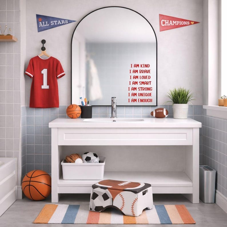 Puede incluir: Un ba&ntilde;o con un tocador blanco, espejo y decoraci&oacute;n deportiva. Una camiseta roja con el n&uacute;mero 1 cuelga de la pared. El espejo tiene las palabras "I AM KIND, BRAVE, LOVED, SMART, STRONG, UNIQUE, ENOUGH" en rojo. Se ven una pelota de baloncesto, un bal&oacute;n de f&uacute;tbol americano y un taburete deportivo.