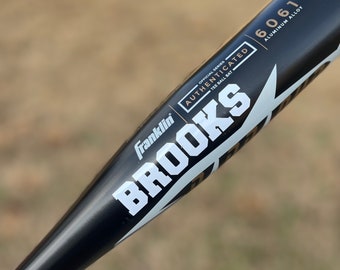 May include: A black baseball bat with white lettering that reads "Franklin Brooks" and "AUTHENTICATED OFFICIAL SERIES TEE BALL BAT" with gold lettering that reads "6061 ALUMINUM ALLOY".