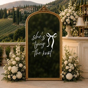 May include: An arched floor mirror with a gold frame featuring the white text "she's tying the knot" and a bow graphic. The mirror is flanked by two large floral arrangements of white roses and greenery, set against a blurred outdoor landscape.