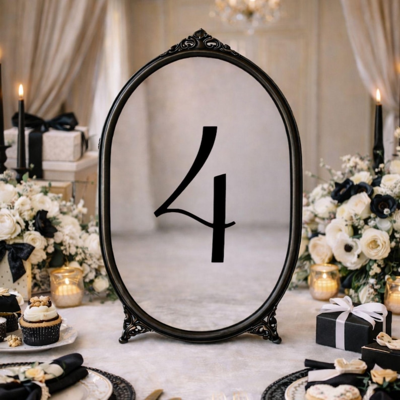 May include: An oval-shaped black frame standing on a base, featuring the number 4 in a black serif font centered on the clear glass surface. The frame has ornate, decorative detailing at the top and bottom, providing a structured display piece.