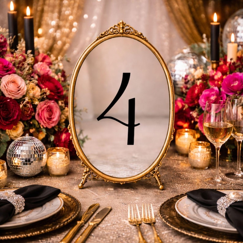 May include: An oval table number sign featuring a gold-toned metal frame with ornate detailing at the top and base. The center of the sign displays the black number 4 in a stylized font. The sign stands upright on a flat surface.