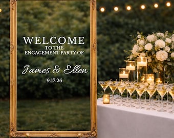 May include: A rectangular gold ornate frame containing a clear sign with white text that reads WELCOME TO THE ENGAGEMENT PARTY OF James & Ellen 9.17.26. The frame is mounted on a stand, positioned outdoors near a table with glassware and candles.