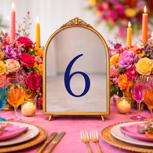 May include: An arched table number sign with a gold-toned metal frame and a reflective surface featuring the dark blue number 6. The sign is positioned upright on a stand, displaying a clear, bold numeral centered on the mirror-like glass panel.