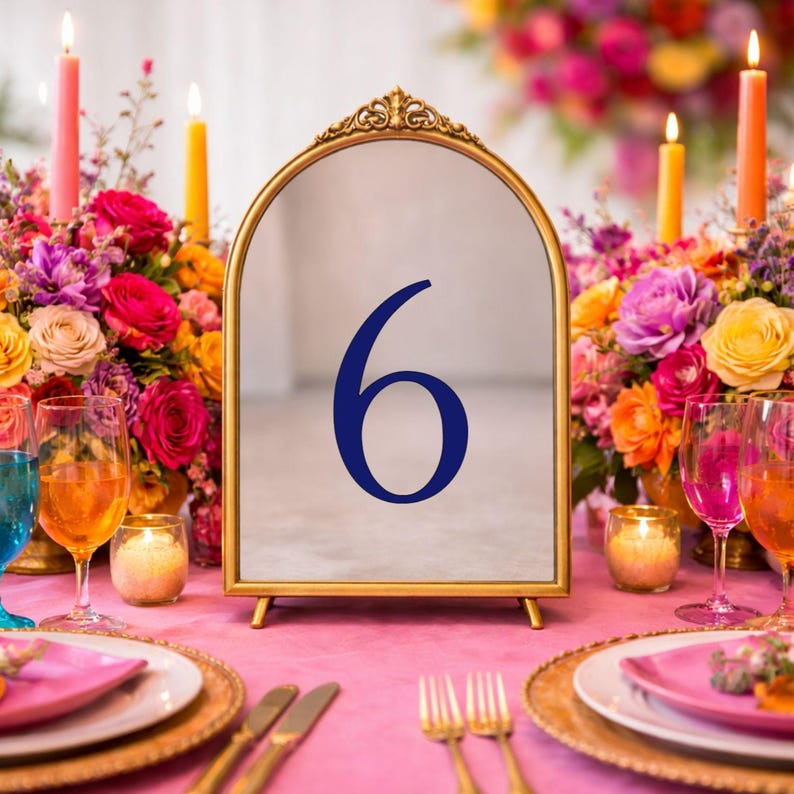 May include: An arched table number sign with a gold-toned metal frame and a reflective surface featuring the dark blue number 6. The sign is positioned upright on a stand, displaying a clear, bold numeral centered on the mirror-like glass panel.