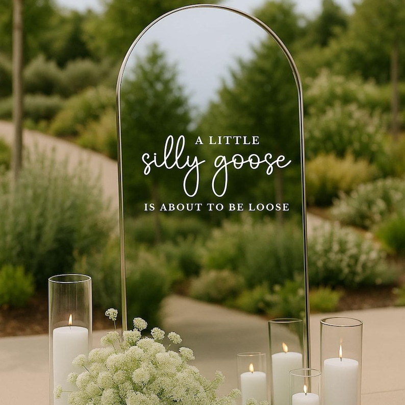 Op de afbeelding: Een gebogen spiegel met een gouden frame toont de tekst "A little silly goose is about to be loose" in witte letters. De spiegel reflecteert een groene buitensc&egrave;ne. Witte kaarsen in glazen cilinders en witte bloemen staan ervoor.