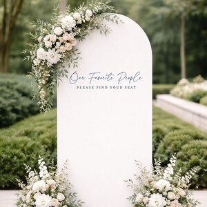 Op de afbeelding: Een wit boogvormig bord met de donkerblauwe tekst Our Favorite People PLEASE FIND YOUR SEAT. Het bord is versierd met een groot arrangement van witte en cr&egrave;mekleurige bloemen en groen blad linksboven en twee kleinere bloemstukken aan de onderkant.