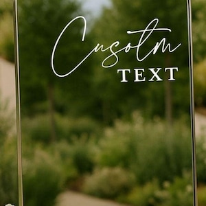 Pu&ograve; includere: Uno specchio ad arco con una sottile cornice metallica che presenta la scritta bianca Custom in corsivo e il testo TEXT in stampatello. Lo specchio riflette una scena esterna sfocata con alberi, fiori bianchi e tre candele alla base.