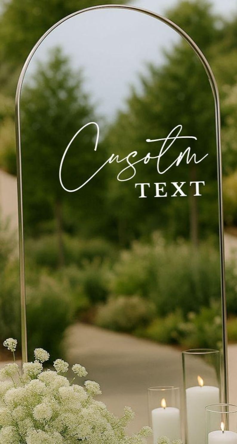 Pu&ograve; includere: Uno specchio ad arco con una sottile cornice metallica che presenta la scritta bianca Custom in corsivo e il testo TEXT in stampatello. Lo specchio riflette una scena esterna sfocata con alberi, fiori bianchi e tre candele alla base.