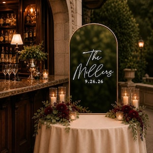 May include: An arched, dark-tinted acrylic wedding sign with a gold metal frame, featuring white script text that reads The Millers and the date 9.26.26. The sign is displayed on a round table covered with a beige cloth, decorated with dark red flowers.
