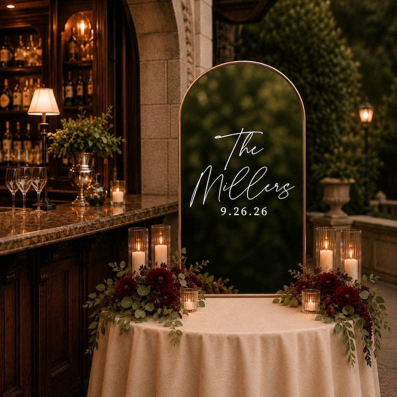 May include: An arched, dark-tinted acrylic wedding sign with a gold metal frame, featuring white script text that reads The Millers and the date 9.26.26. The sign is displayed on a round table covered with a beige cloth, decorated with dark red flowers.