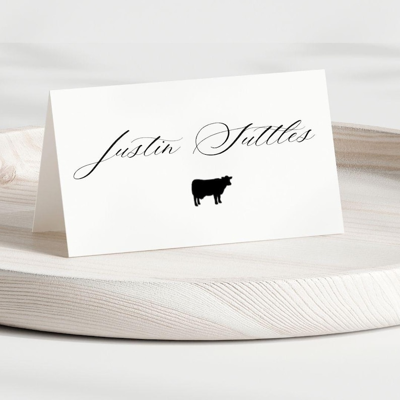 May include: A white folded place card featuring the black printed text Justin Suttles in a cursive script font, positioned above a small black silhouette of a cow. The card is displayed in a tent-fold orientation on a light-colored surface.