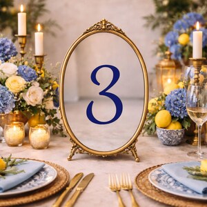 May include: An oval table number sign featuring a gold-toned metal frame with ornate detailing at the top and base. The center of the sign displays the number 3 in a bold, dark blue serif font, positioned upright on a flat surface.