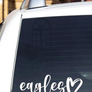 May include: White car with a black tinted back window. The window has a white vinyl decal that says "eagles" with a heart.