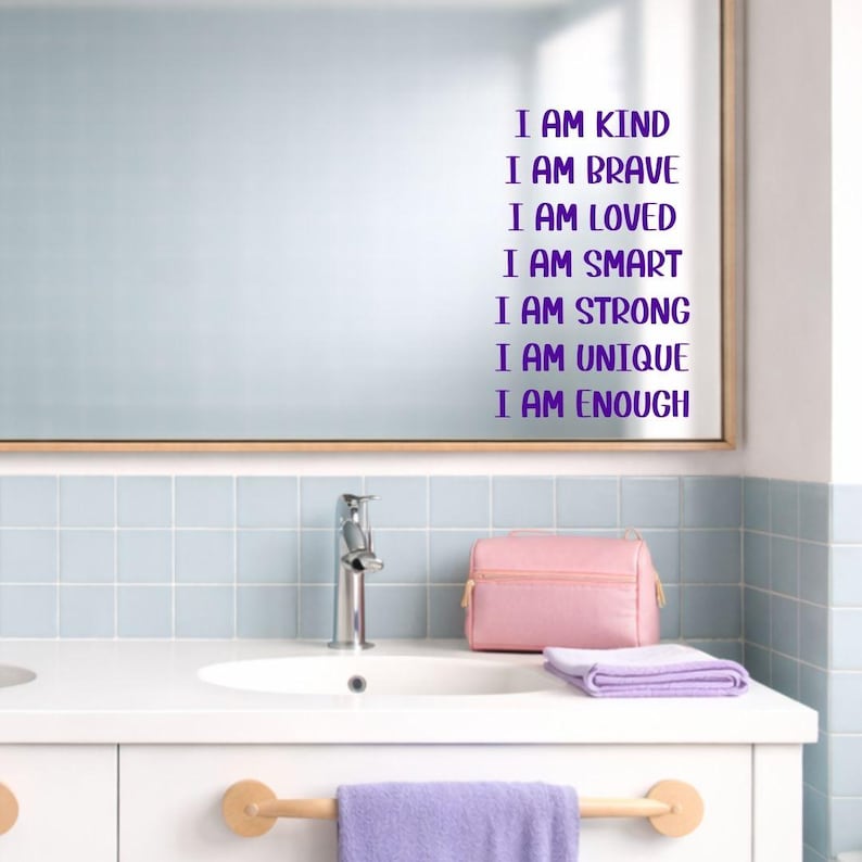 Puede incluir: Un espejo de ba&ntilde;o con texto morado que dice "I AM KIND, I AM BRAVE, I AM LOVED, I AM SMART, I AM STRONG, I AM UNIQUE, I AM ENOUGH." Una bolsa de cosm&eacute;ticos rosa y una toalla morada est&aacute;n en el mostrador.