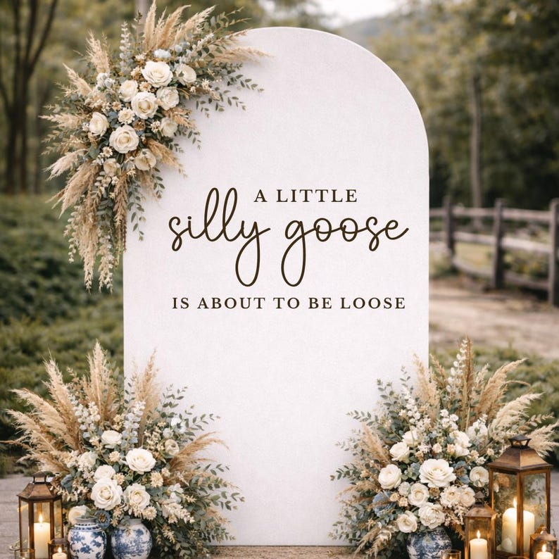 Op de afbeelding: Een wit, boogvormig bord met de tekst "A little silly goose is about to be loose". Het bord is versierd met bloemstukken van witte rozen, pampasgras en groen. Lantaarns en kaarsen flankeren het bord en cre&euml;ren een romantische sfeer.