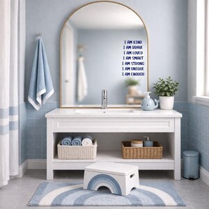 Puede incluir: Un tocador de ba&ntilde;o blanco con un espejo con marco dorado, una alfombra de arco&iacute;ris azul y blanco y un taburete. La pared sobre el tocador tiene el texto "I AM KIND, I AM BRAVE, I AM LOVED, I AM SMART, I AM STRONG, I AM UNIQUE, I AM ENOUGH."